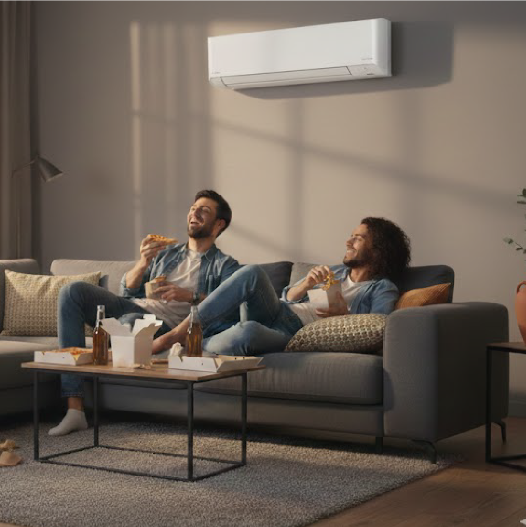Two friends enjoying pizza together on gray sectional sofa in modern living room with wall-mounted air conditioner and large windows.
